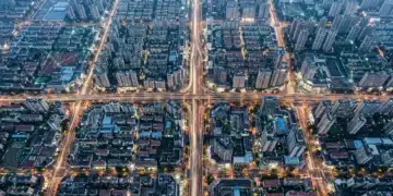 Aerial view of a city at dusk with car light trails forming geometric patterns, overlaid with a faint digital network of green lines, symbolizing interconnected online communities and data streams across urban infrastructure.
