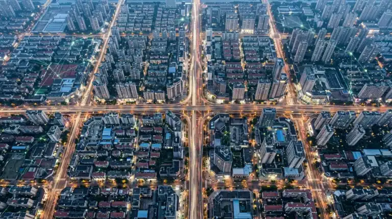 Aerial view of a city at dusk with car light trails forming geometric patterns, overlaid with a faint digital network of green lines, symbolizing interconnected online communities and data streams across urban infrastructure.